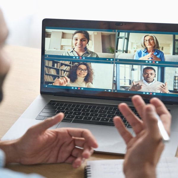 Latin indian businessman having virtual team meeting group call chatting with diverse people in customer support. Video conference call on computer with manager and employees. Over shoulder view.