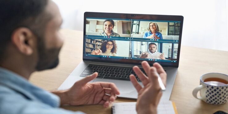 Latin indian businessman having virtual team meeting group call chatting with diverse people in customer support. Video conference call on computer with manager and employees. Over shoulder view.
