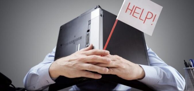 man sitting at desk with a laptop over his head while holding a sign for help