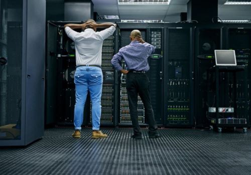 man standing with hands on head next to a nother man staring at servers in  a server room

