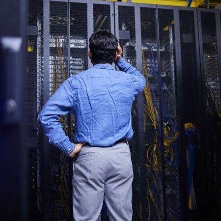 man with his back to the camera scratching his head confused, looking at the computer server