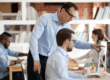 office setting with one man standing over another man at a desk pointing to a monitor