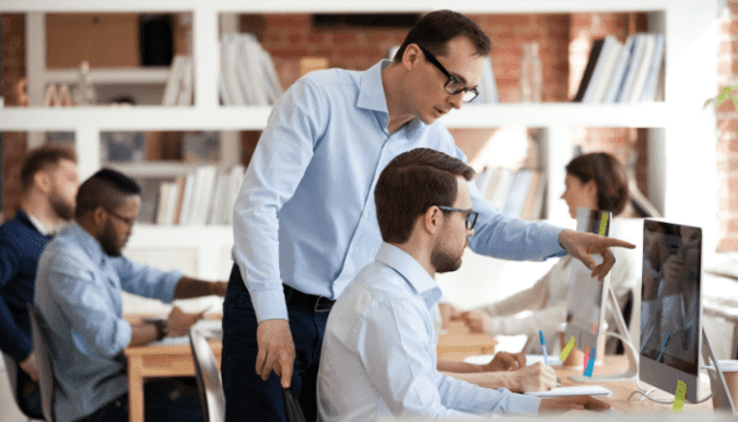 office setting with one man standing over another man at a desk pointing to a monitor